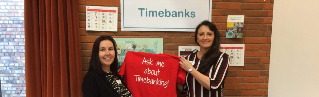 Two women are holding a t shirt that says 'ask me about timebanking'. They are in front of a timebanking display. They're both smiling.