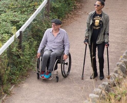 A tall person wearing dark glasses is walking using sticks, next to a man in a wheelchair who is wearing a flat cap and bright blue trainers. They both look happy. They are walking along a path next to a fence, behind which are bushes and shrubs.