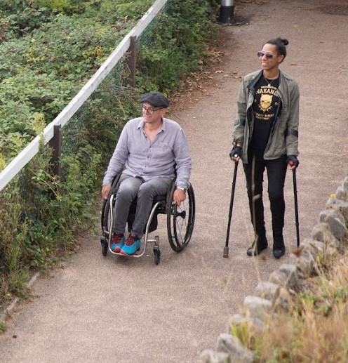 A tall person wearing dark glasses is walking using sticks, next to a man in a wheelchair who is wearing a flat cap and bright blue trainers. They both look happy. They are walking along a path next to a fence, behind which are bushes and shrubs.